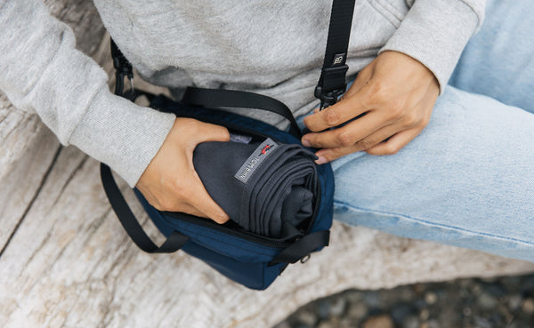 A person sitting on a log  with a Large Shepherd's Wool Utility Cloth in Matter Grey rolled and being stuffed into a Navy Parapack Grab Bag.